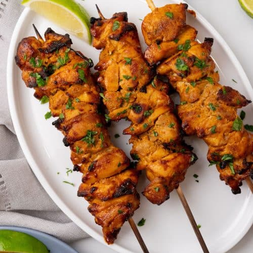 three chicken satay skewers on a white serving plate with fresh cut lime wedges on the side