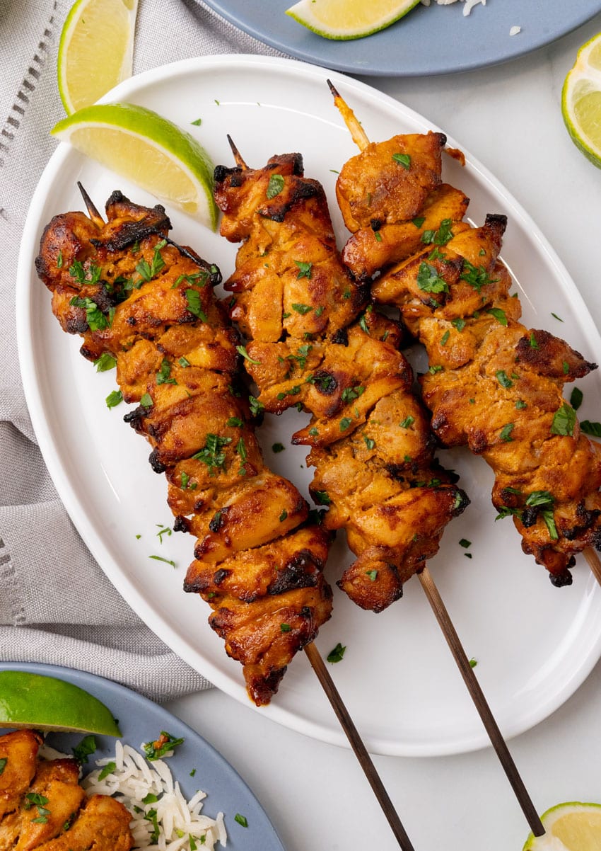 three chicken satay skewers on a white serving plate with fresh cut lime wedges on the side