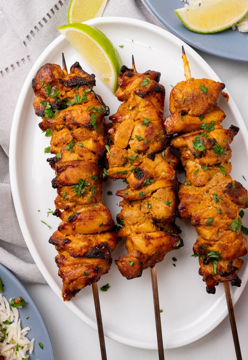 three chicken satay skewers on a white serving plate with fresh cut lime wedges on the side