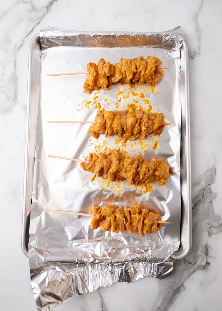 marinaded chicken skewers arranged in a row on an aluminum foil lined baking sheet