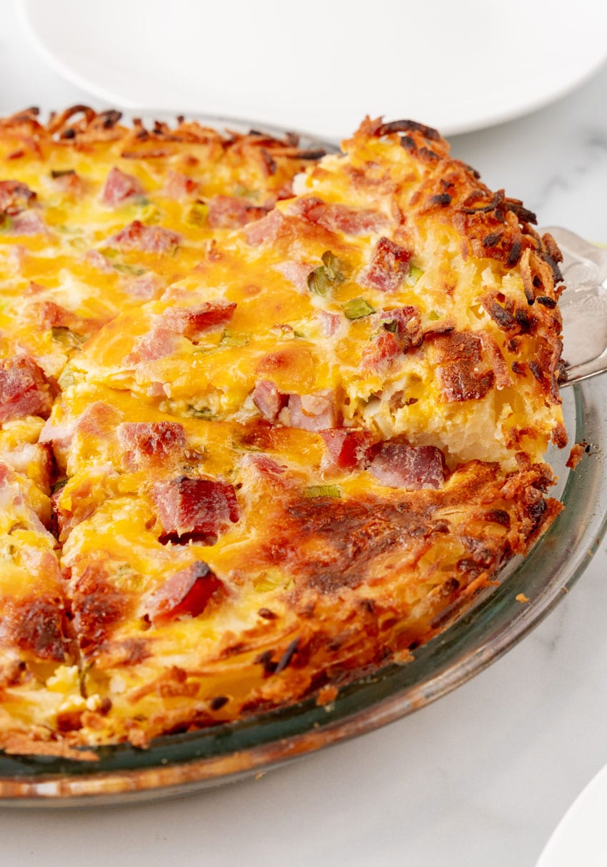a silver spatula lifting a slice of ham and cheese hash brown quiche from a glass pie plate