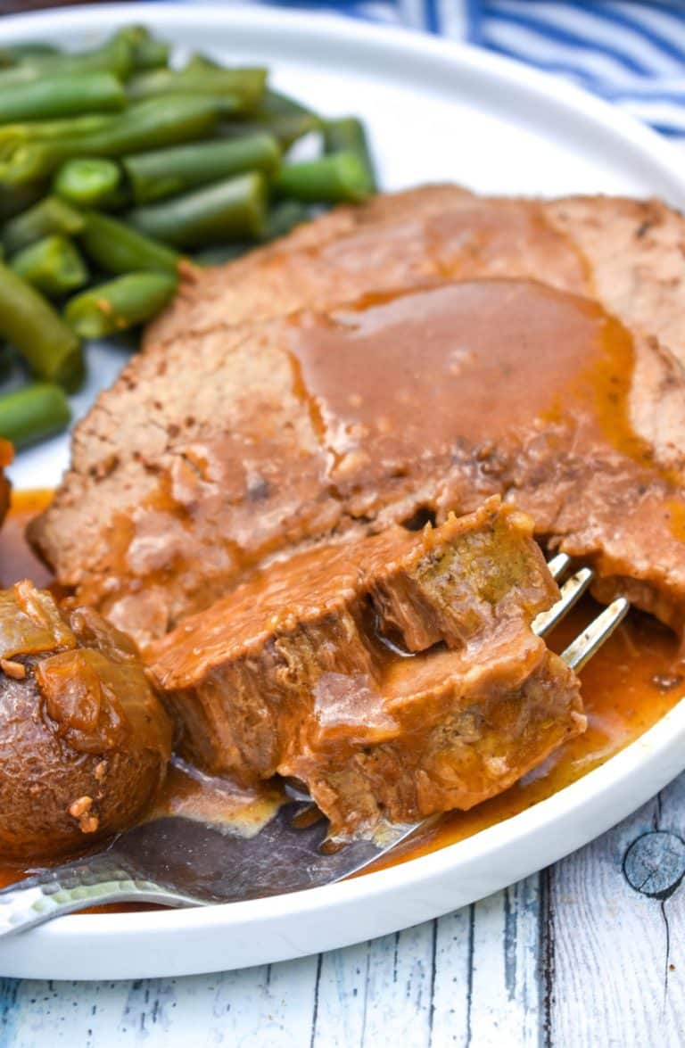 a fork scooping a piece of gravy topped slow cooker eye of round roast off a white plate