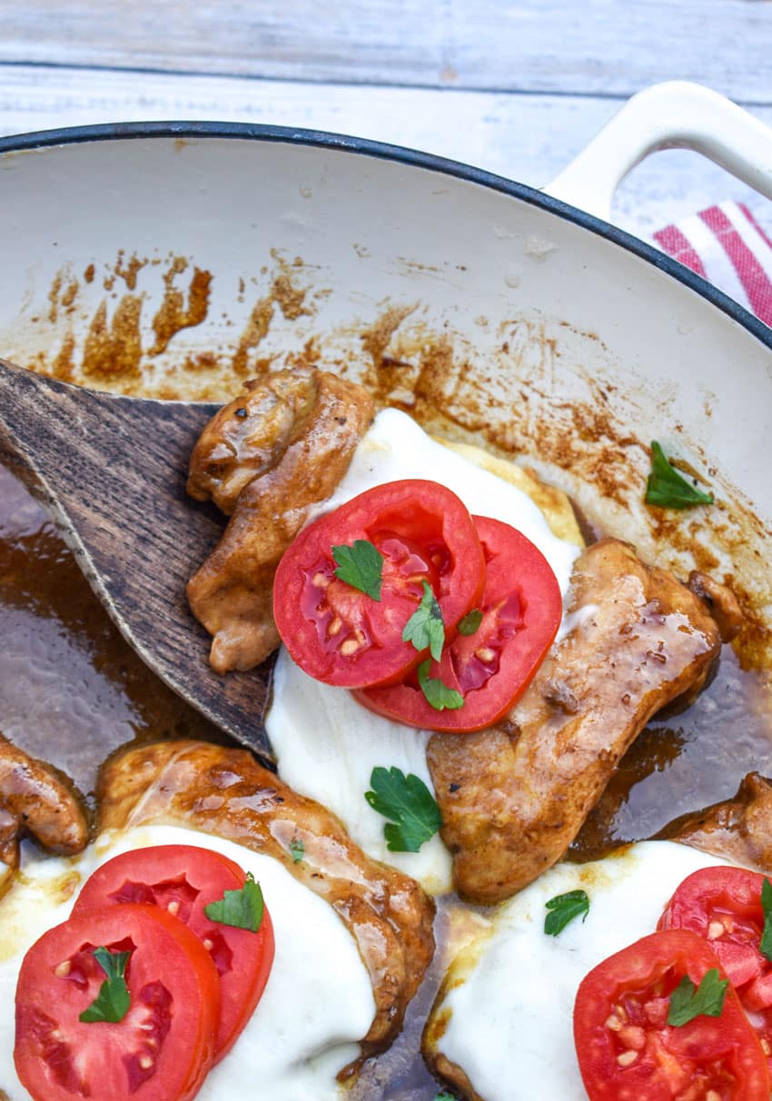 a wooden spatula scooping a slice of balsamic chicken with mozzarella and tomatoes out of a large skillet