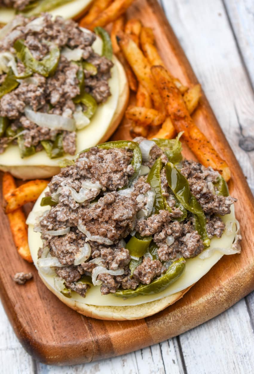 ground beef philly cheesesteak sandwiches surrounded by crispy french fries on a wooden cutting board