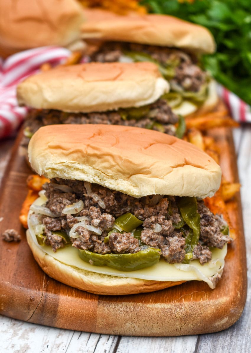 ground beef philly cheesesteak sandwiches surrounded by crispy french fries on a wooden cutting board