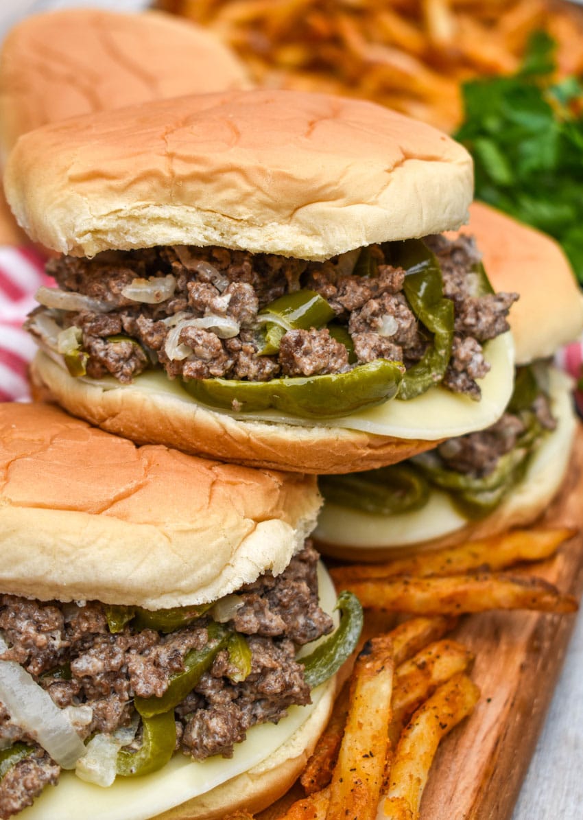 ground beef philly cheesesteak sandwiches surrounded by crispy french fries on a wooden cutting board