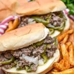 ground beef philly cheesesteak sandwiches surrounded by crispy french fries on a wooden cutting board