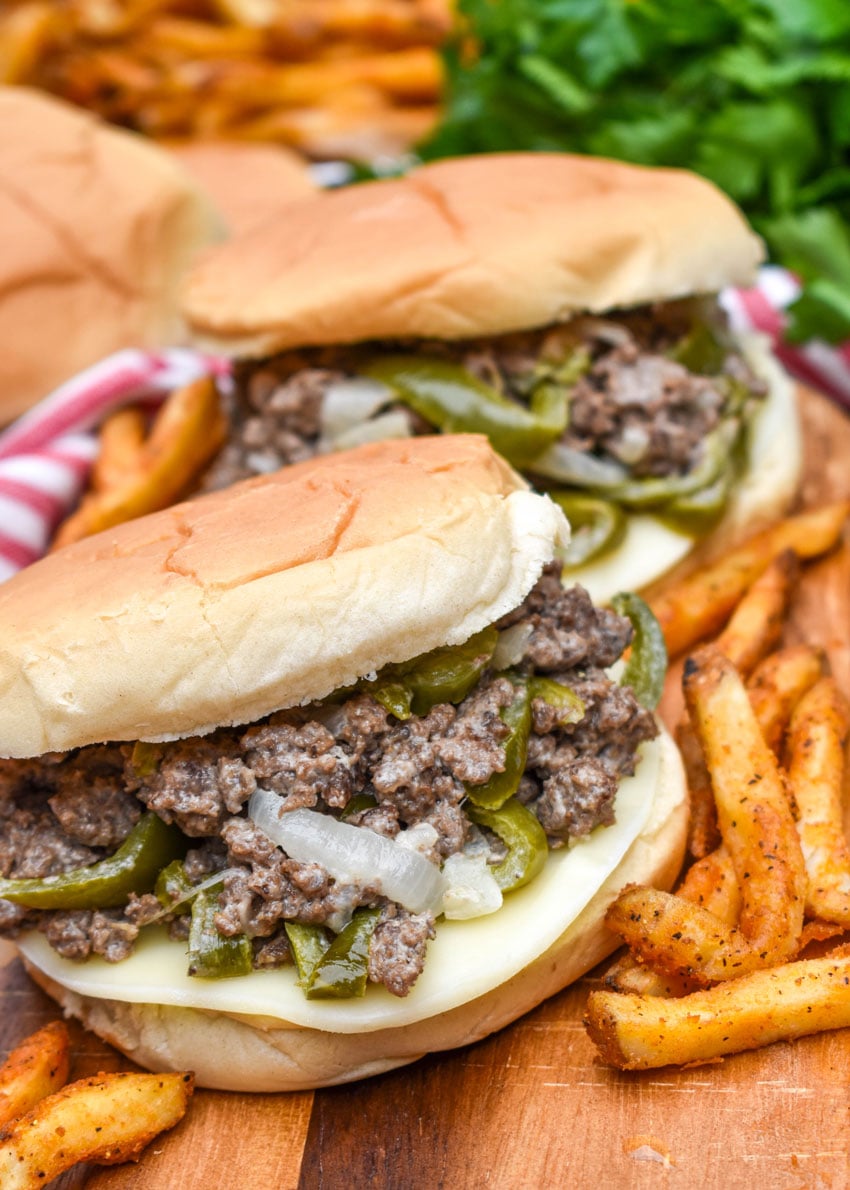 ground beef philly cheesesteak sandwiches surrounded by crispy french fries on a wooden cutting board