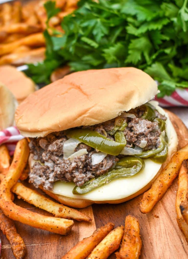 ground beef philly cheesesteak sandwiches surrounded by crispy french fries on a wooden cutting board