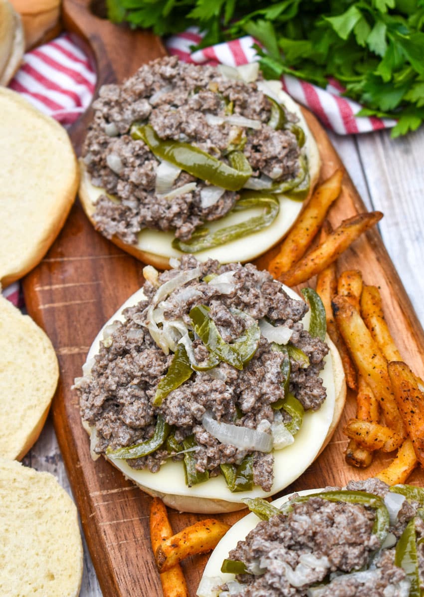 ground beef philly cheesesteak sandwiches surrounded by crispy french fries on a wooden cutting board