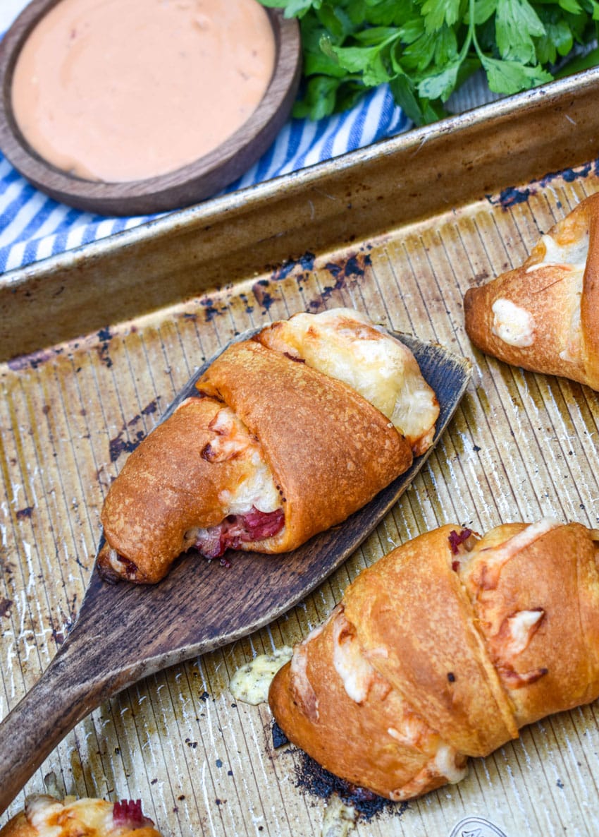 a wooden spatula lifting a baked reuben crescent roll off a metal baking sheet pan