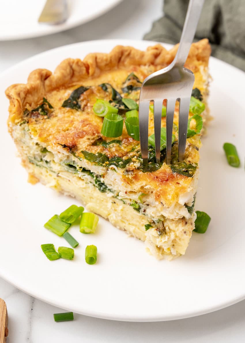 a slice of crab quiche on a small white plate with a silver fork resting on the side