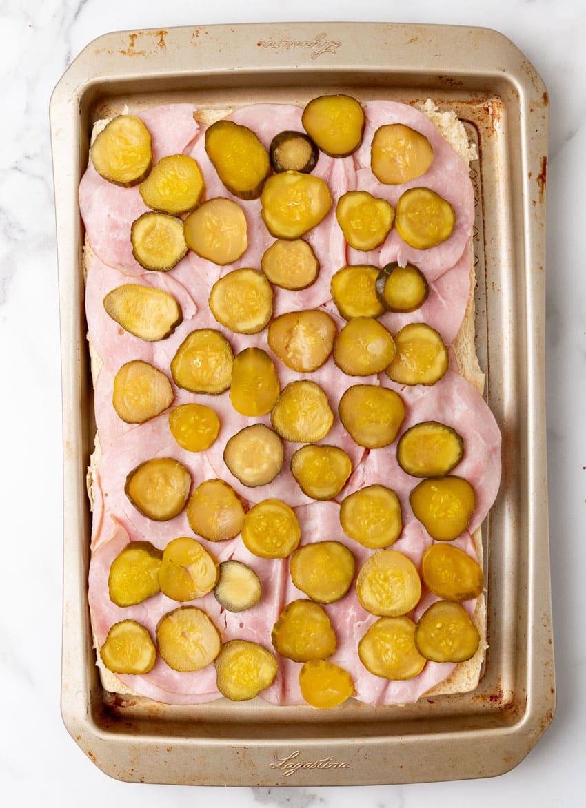 sliced hawaiian rolls topped with slices of ham and pickles on a rimmed baking sheet