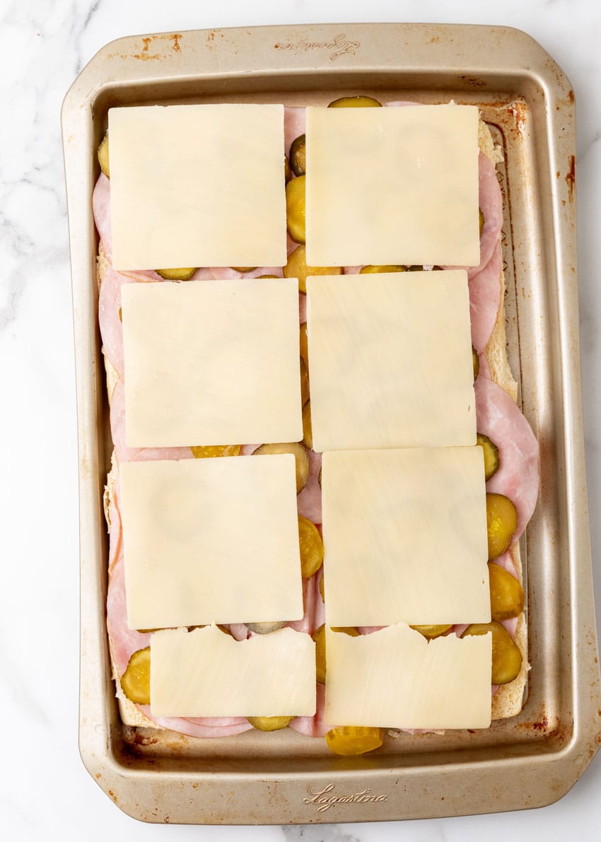 sliced hawaiian rolls topped with slices of ham, pickles, and swiss cheese on a rimmed baking sheet