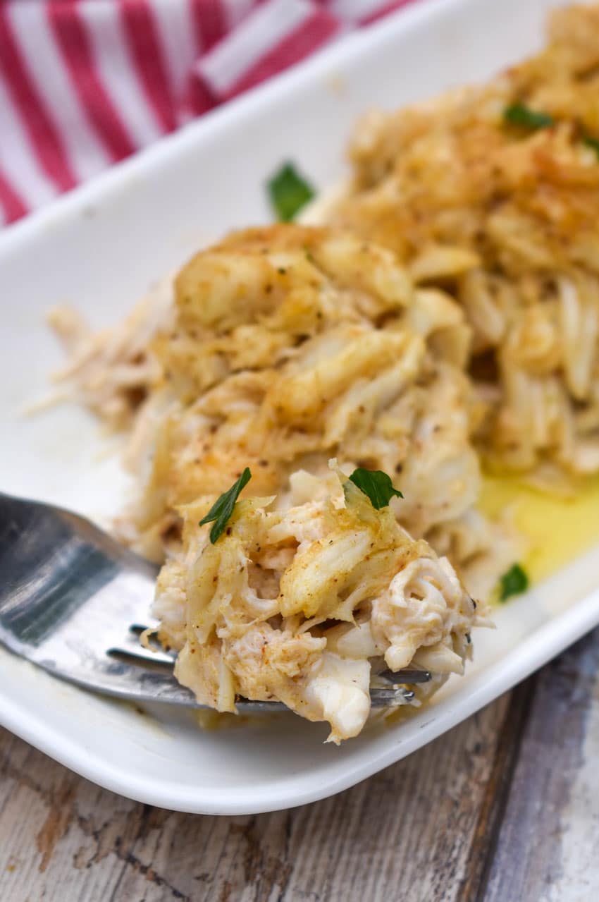 a silver fork holding a piece of a baked lump crab cake resting on the edge of a white serving plate