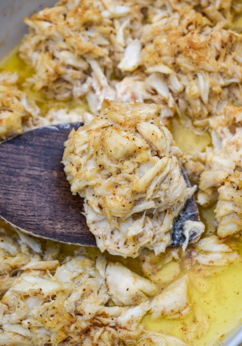 a wooden spatula lifting a baked lump crab cake out of a butter filled white baking dish