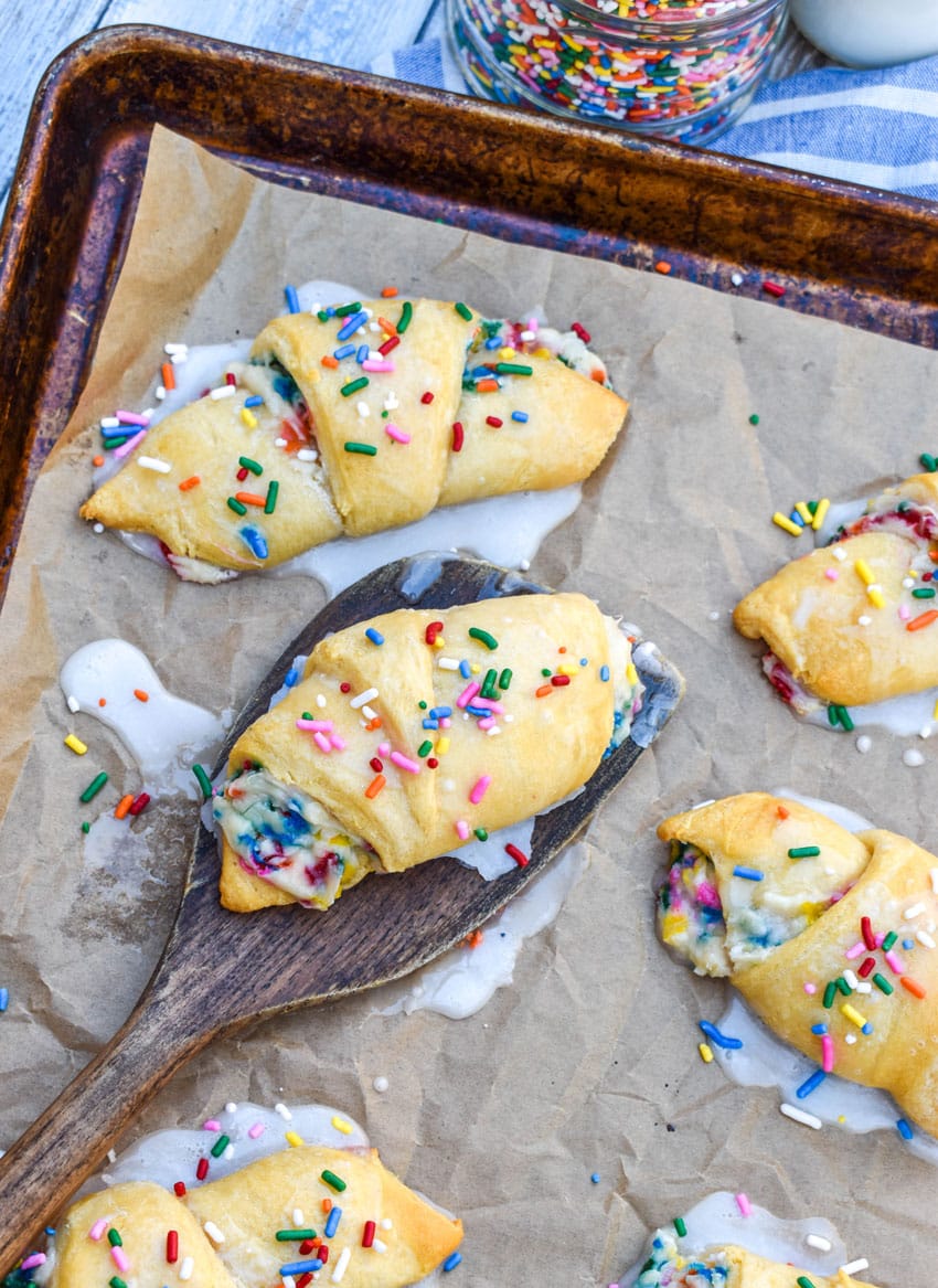 a wooden spatula lifting a funfetti cheesecake stuffed crescent roll off a parchment paper lined baking sheet