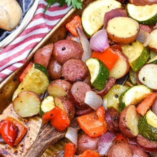 a wooden spatula scooping slices of smoked sausage and roasted vegetables off a metal sheet pan