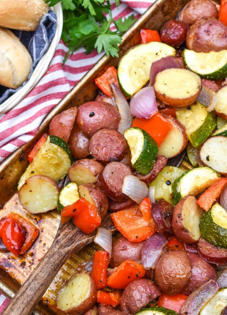a wooden spatula scooping slices of smoked sausage and roasted vegetables off a metal sheet pan