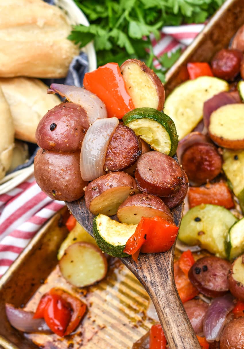 a wooden spatula holding up slices of smoked sausage and roasted vegetables