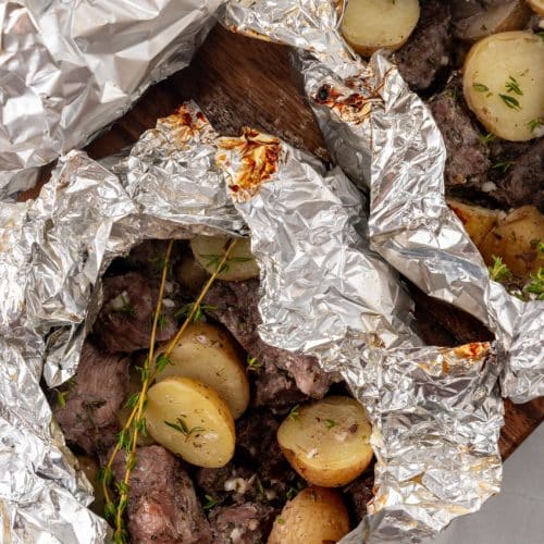 an opened garlic butter grilled steal and potato foil packet resting on a wooden cutting board