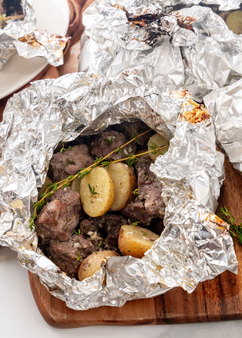 an opened garlic butter grilled steal and potato foil packet resting on a wooden cutting board