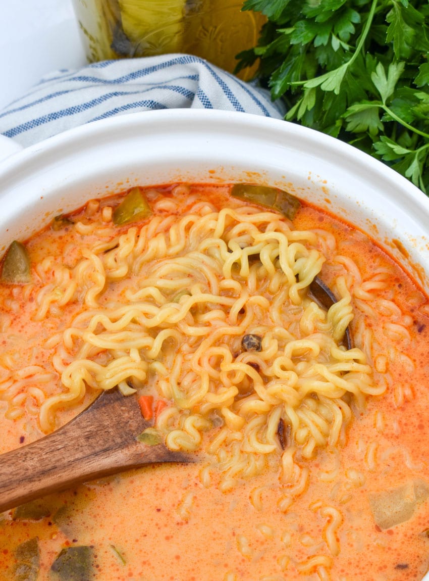 a wooden ladle scooping cheesy ramen noodles out of a small white soup pot