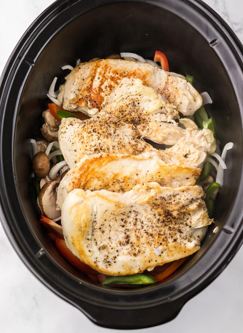 seared seasoned chicken breasts over sauteed peppers and onions in the black bowl of a slow cooker