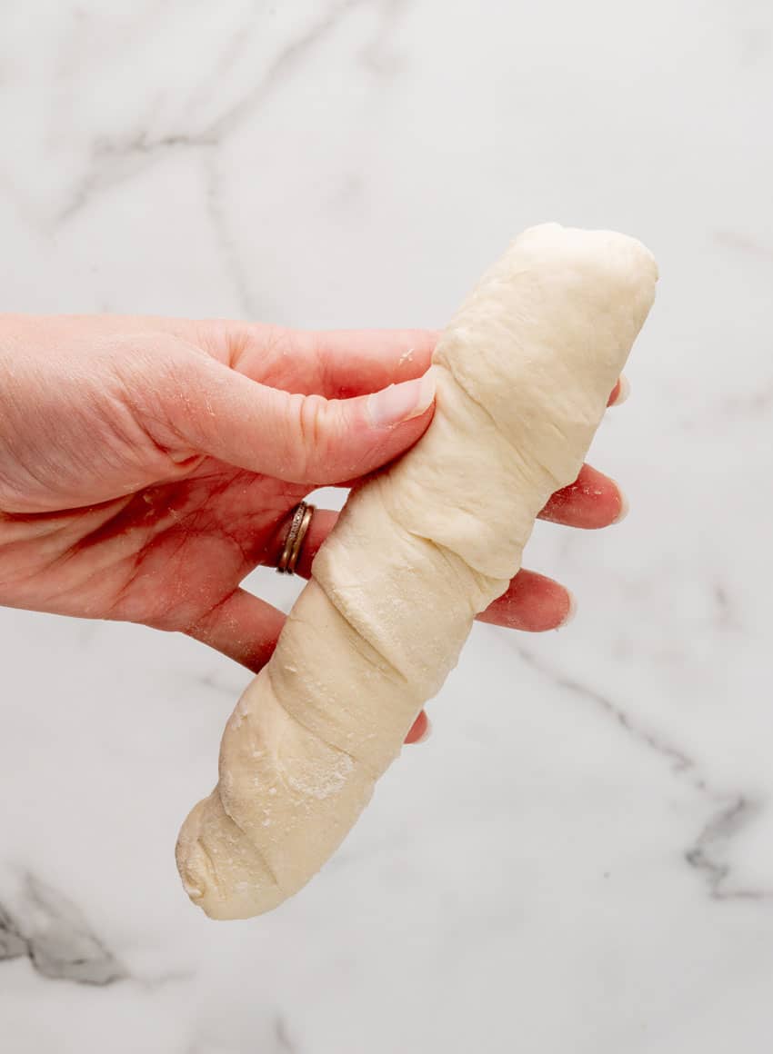 a hand holding an uncooked dough wrapped homemade pretzel dog