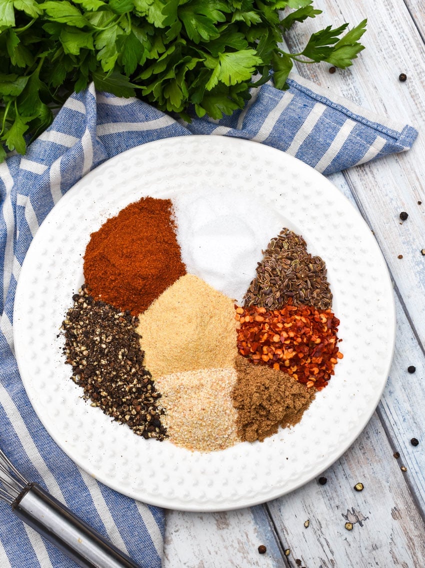 an overhead image showing all of the measured spices needed for making copycat montreal steak seasoning on a small white plate