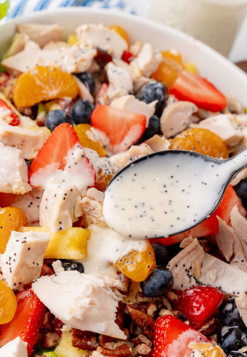 a black spoon drizzling homemade poppyseed dressing over a chicken salad in a white serving bowl