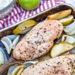a wooden spatula lifting a piece of baked chicken surrounded by roasted apples and onions of a metal sheet pan