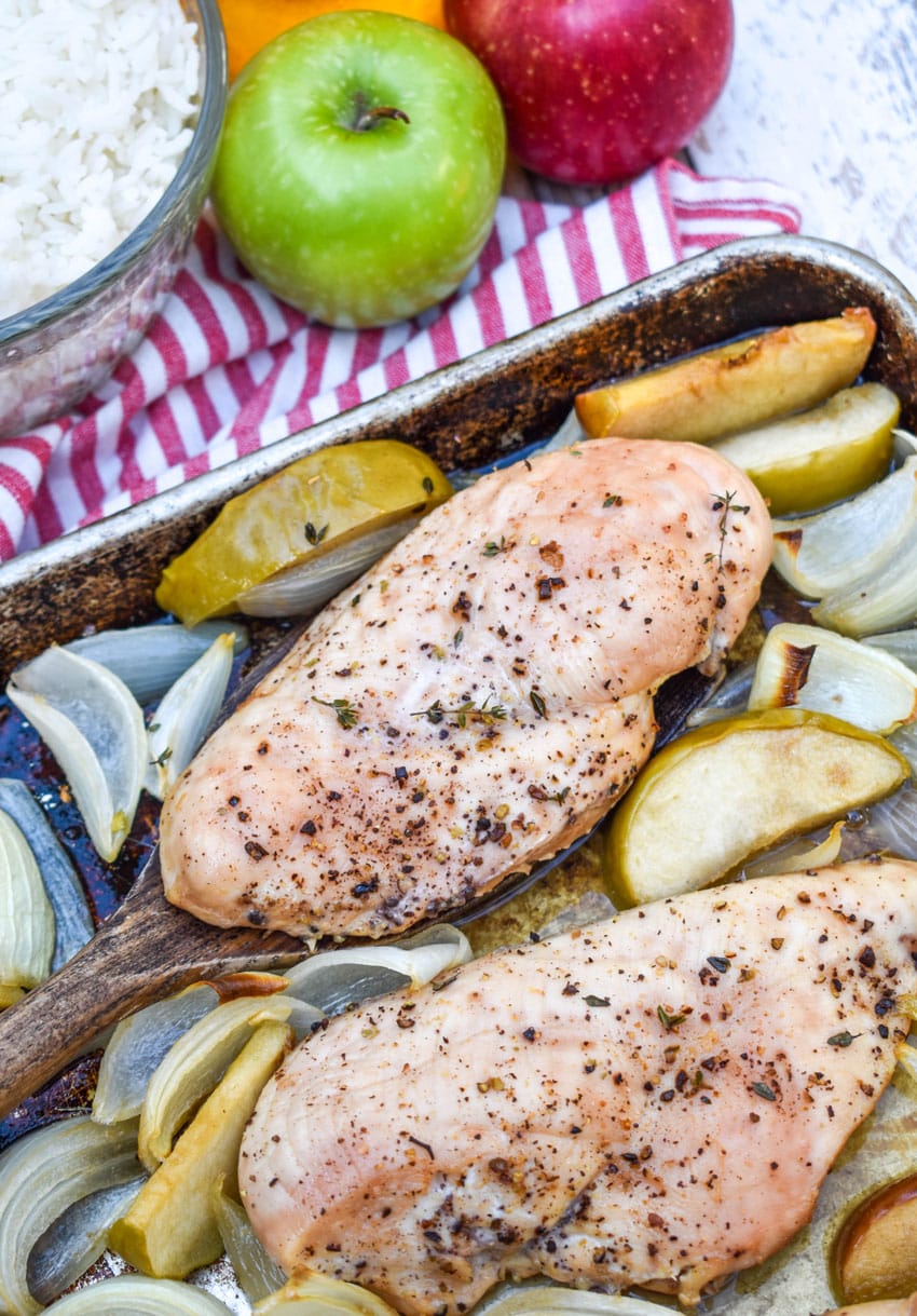 a wooden spatula lifting a piece of baked chicken surrounded by roasted apples and onions of a metal sheet pan