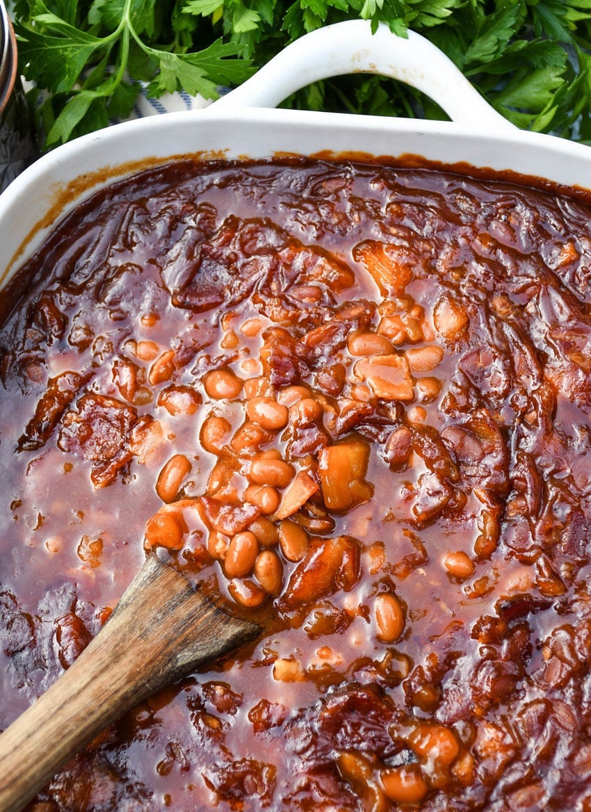 a wooden spoon scooping homemade apple pie baked beans out of a white casserole dish