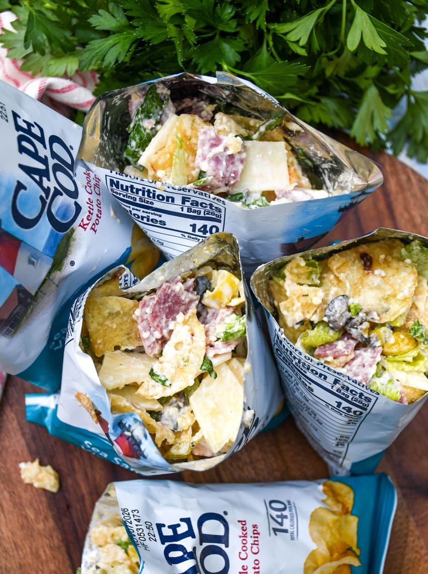 a group of prepared chip bag sandwiches on a wooden serving board with fresh herbs in the background