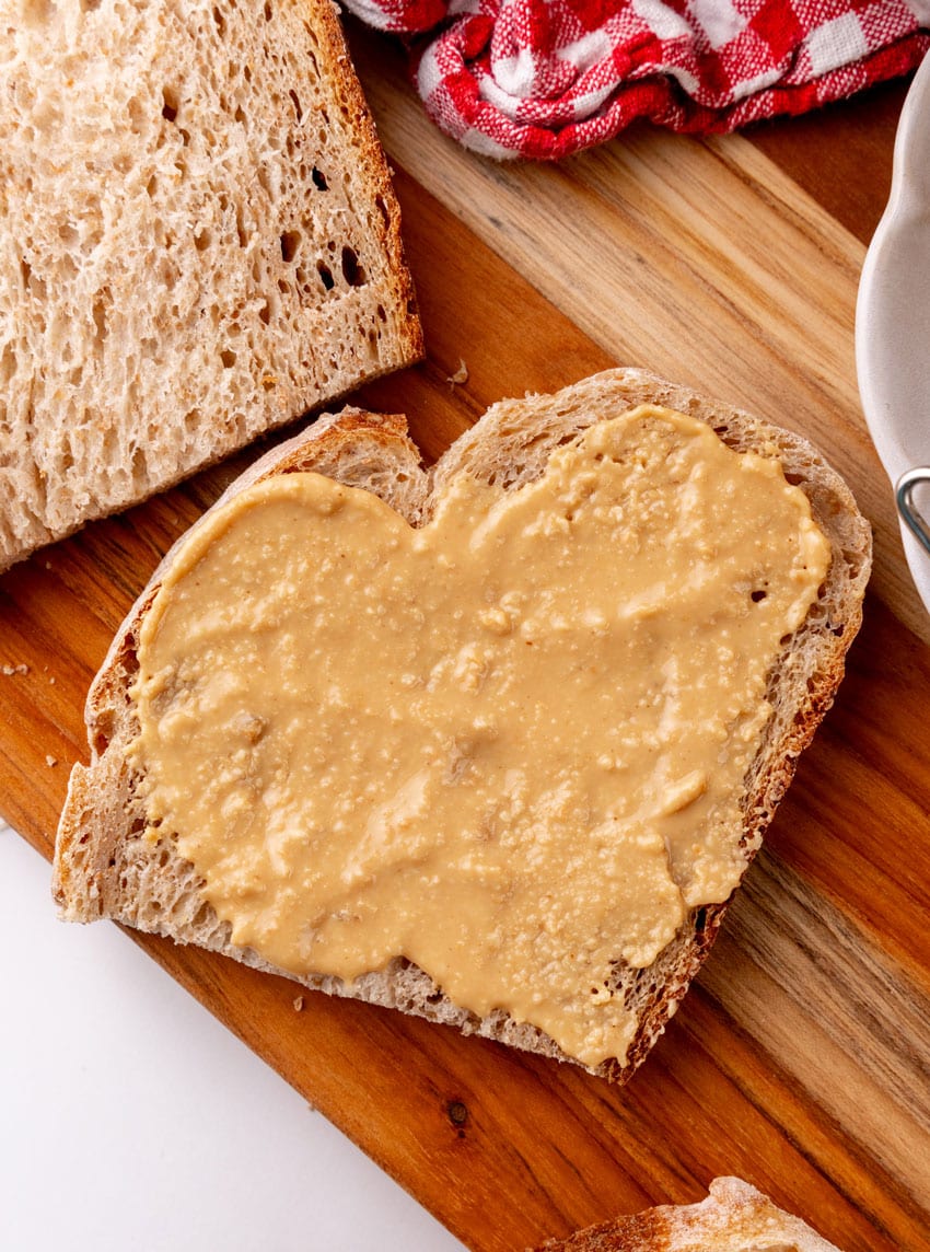 a slice of soft wheat sandwich bread spread with homemade peanut butter resting on a wooden serving board