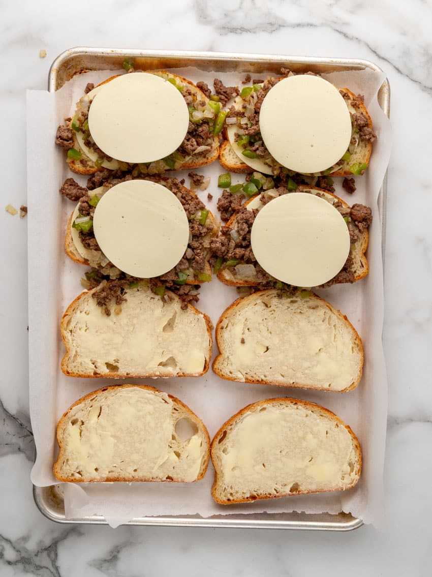 sauteed ground beef with peppers and onions topped with provolone cheese on buttered slices of sourdough bread on a parchment paper lined baking tray