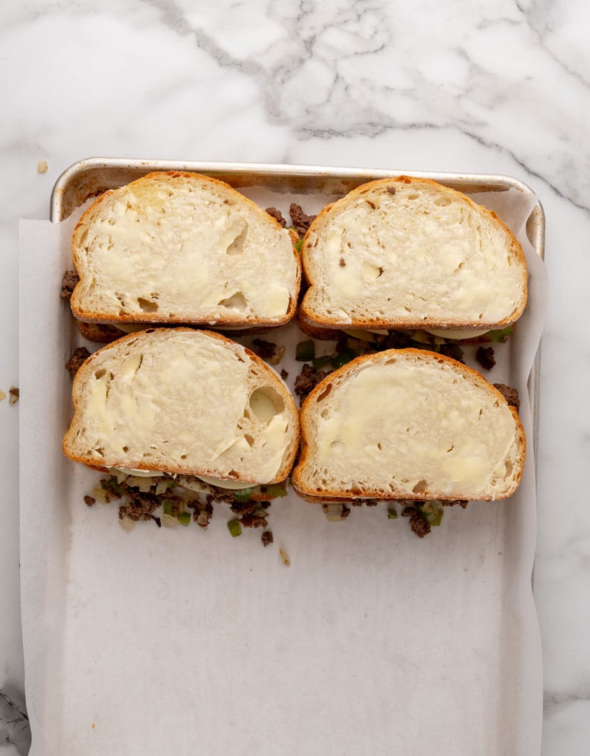 prepped ground beef philly cheesesteak grilled cheese sandwiches on a parchment paper lined baking sheet 