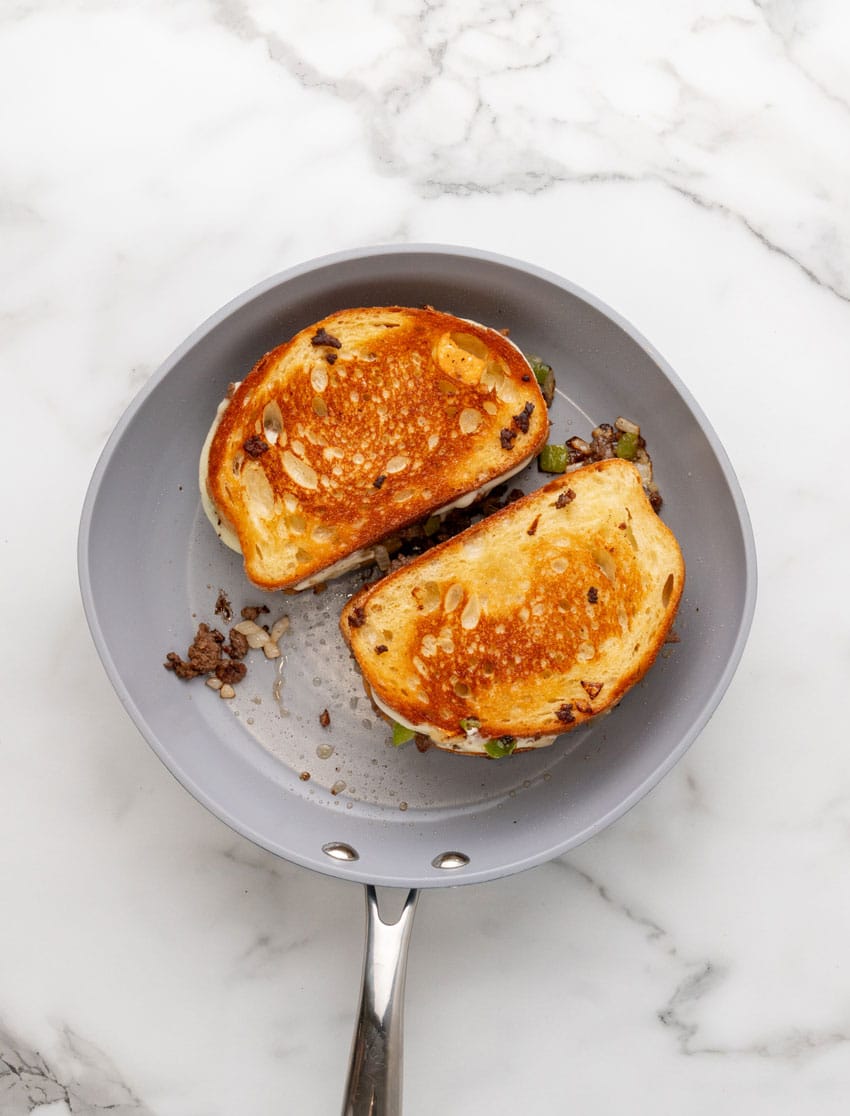 two ground beef philly cheesesteak grilled cheese sandwiches cooking in a gray skillet