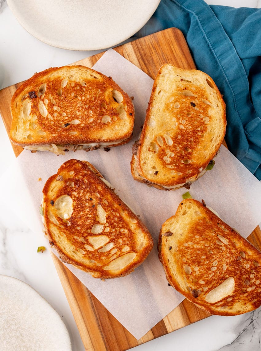ground beef philly cheesesteak grilled cheese sandwiches on a parchment paper lined wooden cutting board