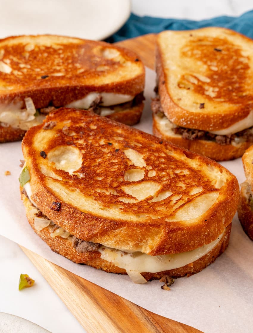 a close up of ground beef philly cheesesteak grilled cheese sandwiches on a parchment paper lined wooden cutting board