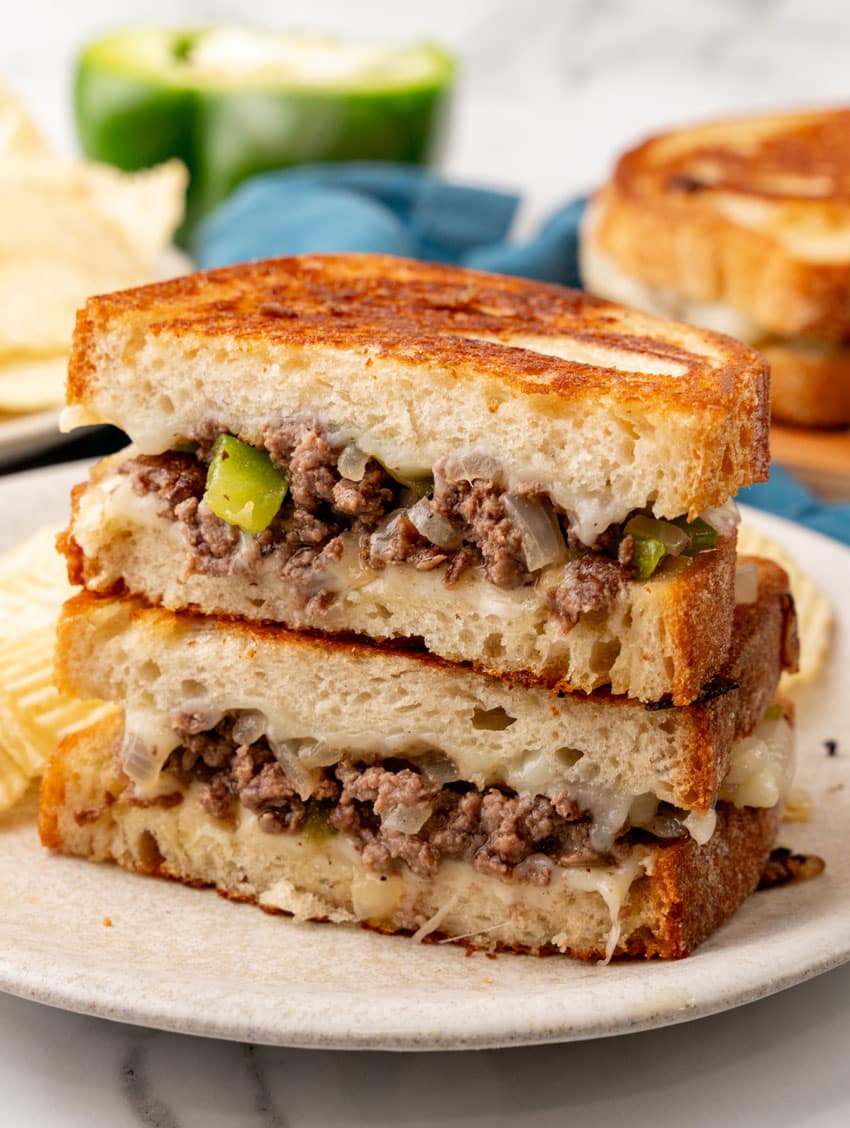 two halves of a philly cheesesteak grilled cheese sandwich stacked on a white plate with potato chips on the side