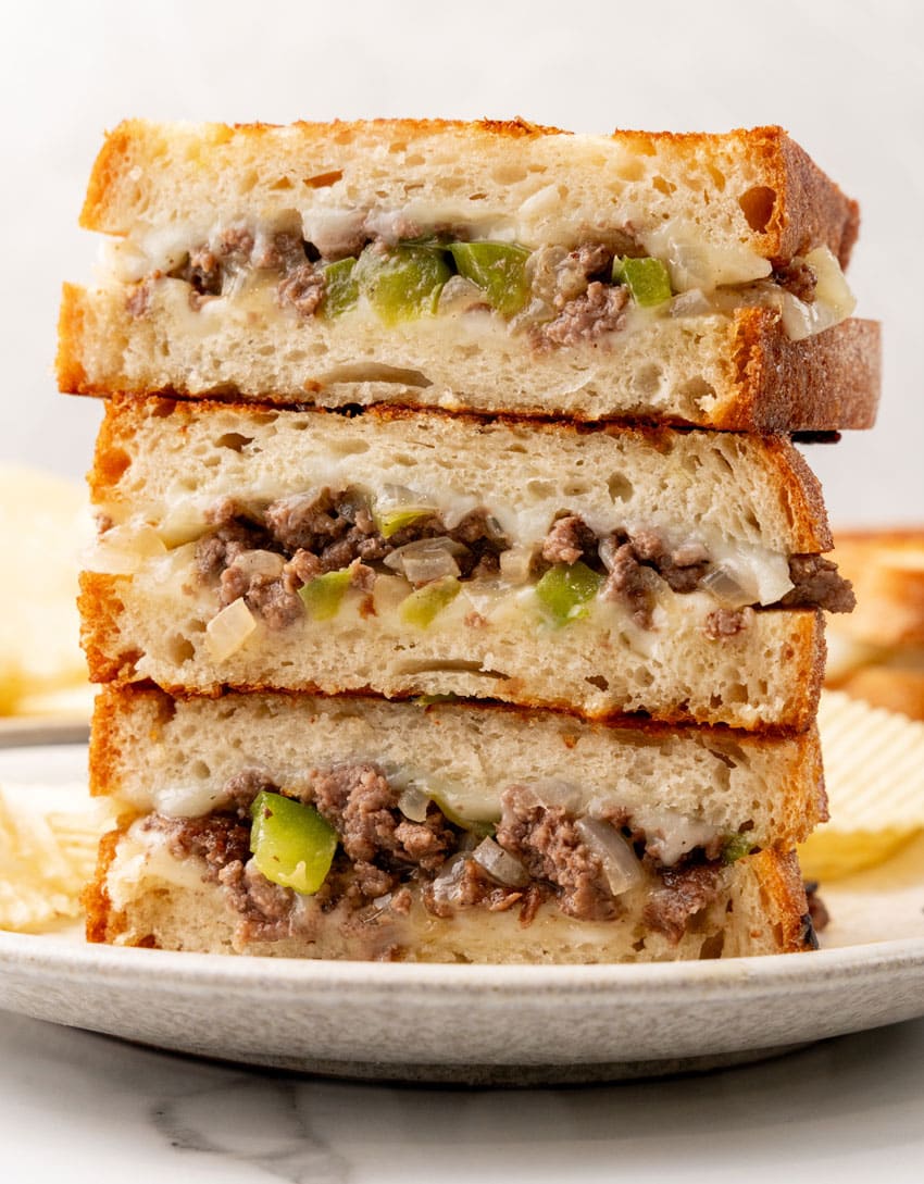 three halves of philly cheesesteak grilled cheese sandwiches stacked on a white plate with potato chips on the side