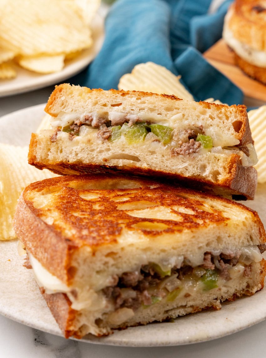 two halves of a philly cheesesteak grilled cheese sandwich stacked on a white plate with potato chips on the side