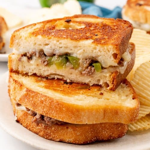 two halves of a philly cheesesteak grilled cheese sandwich stacked on a white plate with potato chips on the side