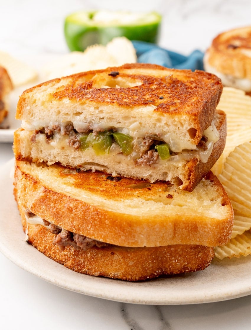 two halves of a philly cheesesteak grilled cheese sandwich stacked on a white plate with potato chips on the side