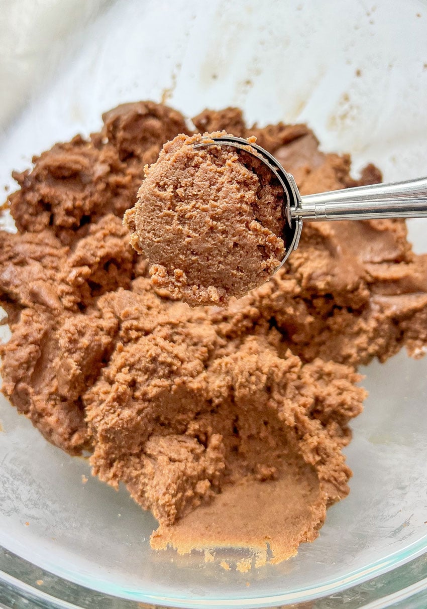 A SMALL COOKIE SCOOP SCOOPING BROWNIE CHEESECAKE TRUFFLE MIX OUT OF A GLASS MIXING BOWL