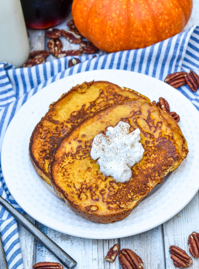 two slices of pumpkin french toast topped with syrup and a dollop of whipped cream on a small white plate with a silver fork resting on the edge