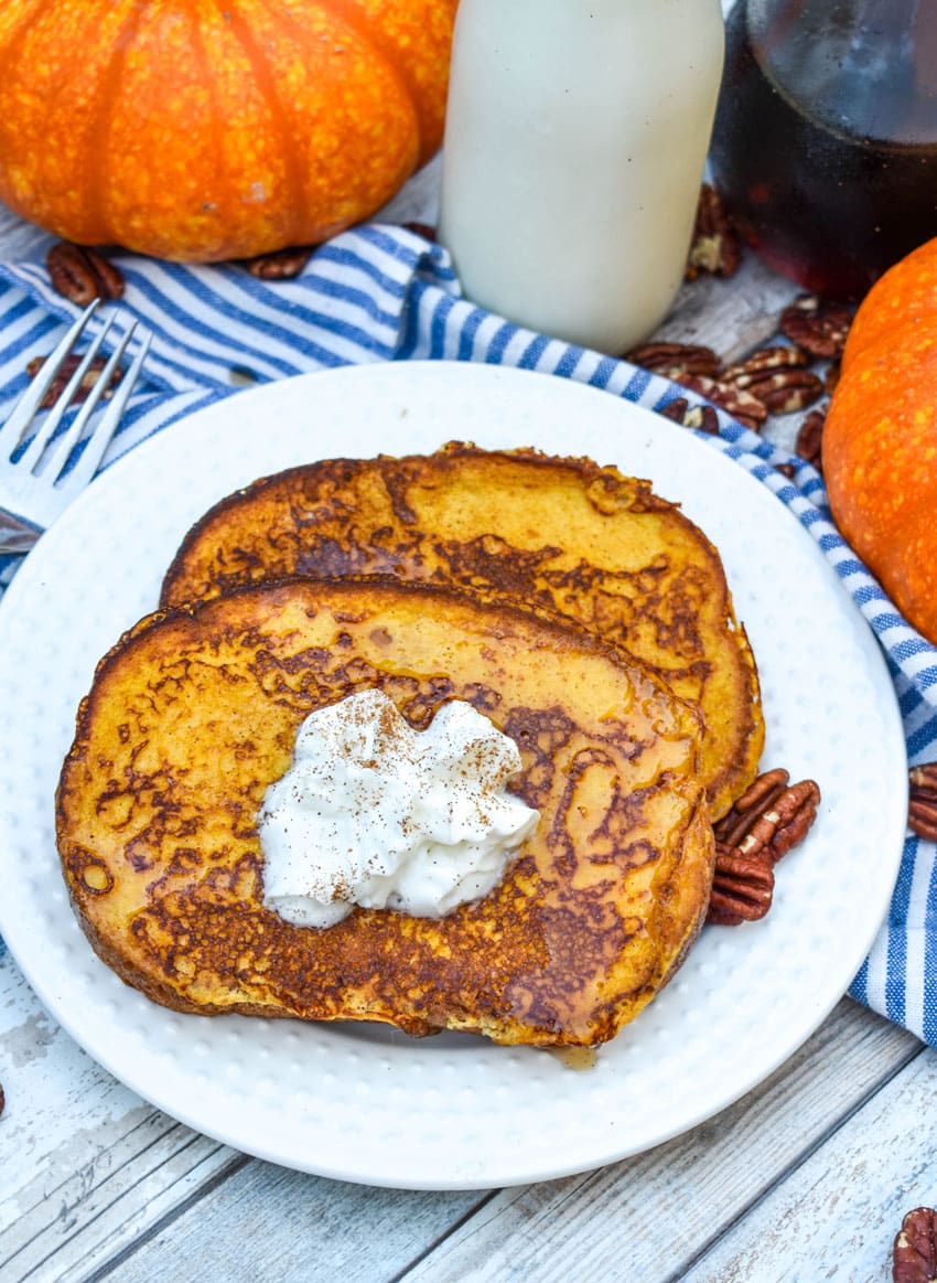 two slices of pumpkin french toast topped with syrup and a dollop of whipped cream on a small white plate with a silver fork resting on the edge