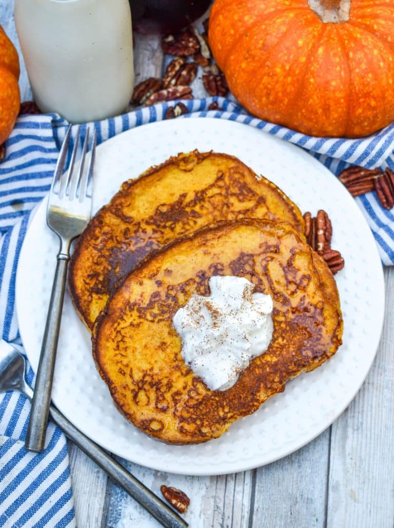 two slices of pumpkin french toast topped with syrup and a dollop of whipped cream on a small white plate with a silver fork resting on the edge